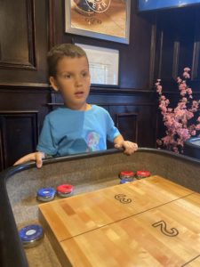 Image of a boy standing at the end of shuffle board table. He appears to be intently watching where his puck has gone.